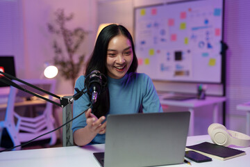 Asian woman smiles while recording a podcast in her home studio, using a professional microphone...