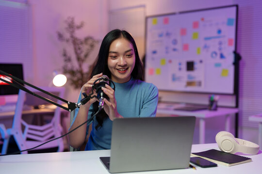 Young content creator recording a podcast episode using professional microphone and laptop in her home studio, illuminated by colorful led lights