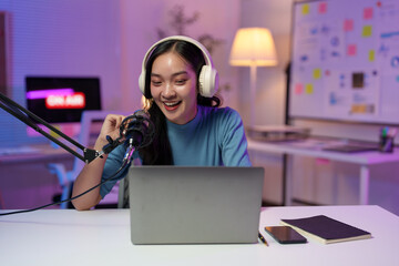 Young cheerful asian woman radio host wearing headphones recording podcast using microphone and laptop in home studio, gesturing while talking during online broadcast