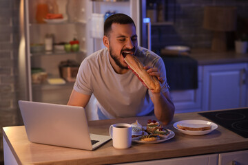 Hungry young man with laptop and cup of tea eating sandwich in kitchen at night