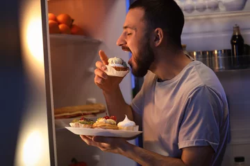 Fotobehang Hungry young man eating cupcake near refrigerator in kitchen at night © Pixel-Shot