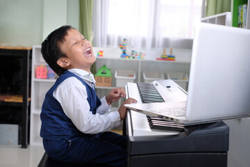 An adorable Asian child is laughing while attending an online piano class with a laptop.