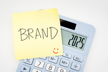 BRAND text on sticky note with light blue Calculator. Build your Brand in 2025. Brand building, success, business. Calculator and forecast for finances in 2025.