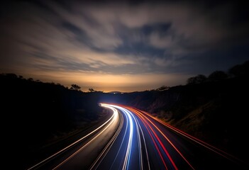 Fototapeta premium highway at night with long exposure light trails of red and white from vehicles, surrounded by a forested landscape under a deep blue sky with streaking clouds. 