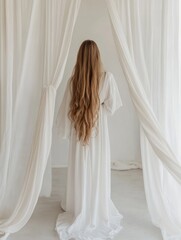 Elegant young woman with flowing long hair in a white robe standing beneath sheer white drapes, soft morning light, spacious empty backdrop for text, studio setting.