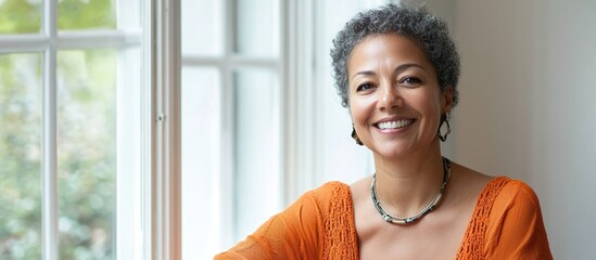 Smiling biracial middle-aged woman in casual orange shirt by sunlit window at home, showcasing natural beauty with ample empty space for text.