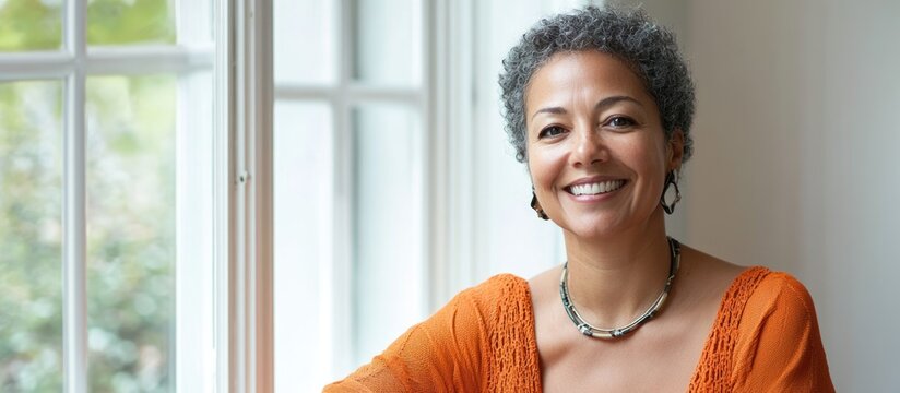 Smiling biracial middle-aged woman in casual orange shirt by sunlit window at home, showcasing natural beauty with ample empty space for text.