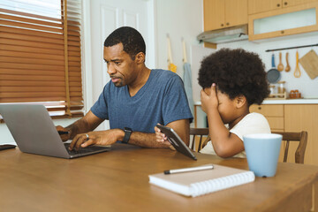Black African American Father working on laptop while son uses tablet at kitchen table, showing multitasking and shared family time with technology.