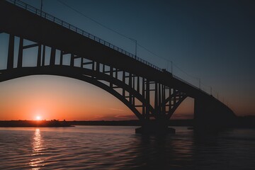 Naklejka premium Silhouetted steel arch bridge at sunset over a calm river. The bridges structure is emphasized against the vibrant sky. Strong lines and contrasting colors create a dramatic image.