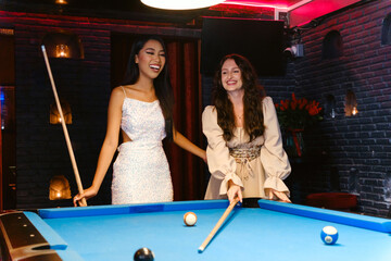 Two women enjoy a fun evening at a billiard table, laughing and playing pool in a stylish, dimly...
