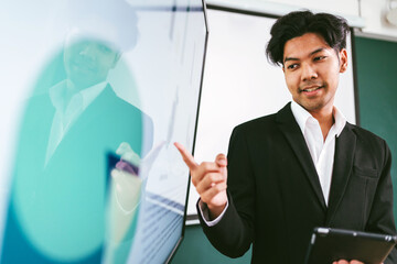 A confident young professional presents information using a digital display, engaging an audience with clear communication and a polished appearance. Asian businessman leader role at the meeting.