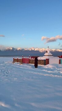 Vertical video of a girl tourist taking a photo of a white Buddhist stupa or suburgan against the backdrop of the snow-capped mountains of the Eastern Sayan Mountains at winter sunset