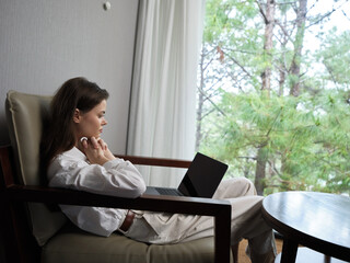 Young woman in cozy attire, enjoying work on laptop in serene indoor space with nature view, reflecting peace and productivity