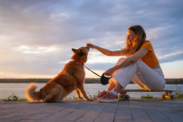 Young woman enjoys a relaxing evening by the river, sitting on her skateboard and feeding her karelian bear dog a treat as the sun sets, creating a warm and peaceful atmosphere © wifesun