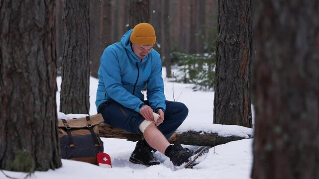 Wounded hiker carefully bandaging injured knee in snow-covered forest, applying first aid during challenging winter trek with medical kit