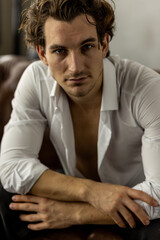 Obraz premium Young sexy handsome fit stylish man in a white shirt looking at the camera sitting on a chair in the studio. Portrait in a studio, casual clothes, modern style