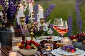 Elegant gorgeous wedding table decor or romantic dinner arrangement outdoors in blooming field. Purple lupine flowers, candles, fruits and wine, wooden vintage furniture. Sunset, summer, golden hour