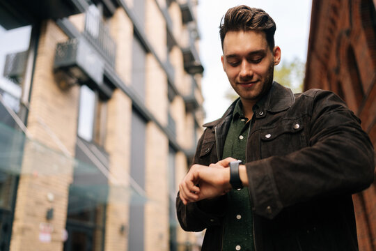 Low-angle portrait of fashionable young man in brown leather jacket checking smartwatch outside modern building, possibly during workday commute or break. Concept of modern successful city lifestyle.