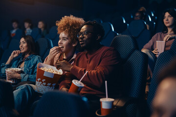 Happy multiracial couple eating popcorn during movie projection at cinema.