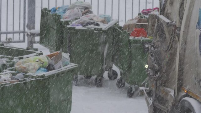 Snow-covered day with garbage truck emptying row of green dumpsters in snowy city. Removing municipal solid waste from residential collection site with garbage truck in snowfall.