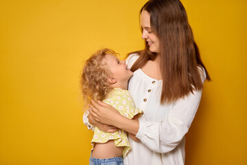 A smiling mother holds her cheerful child in her arms, both enjoying a playful interaction. The bright yellow backdrop enhances their warm connection and happiness