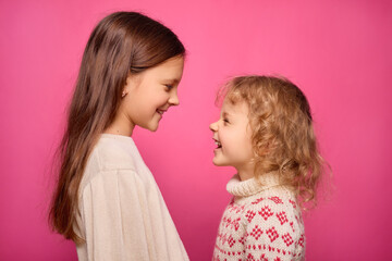 Two young girls stand close together, smiling and looking into each other's eyes, radiating joy and friendship in front of a vibrant pink backdrop