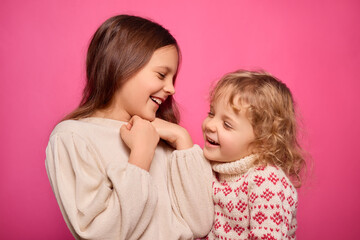 Two girls stand close together, smiling and laughing with joy. One girl has long hair and a light sweater, while the other has curly hair and a patterned sweater