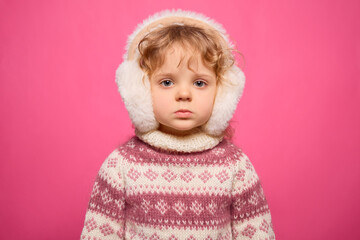 A young child dressed warmly in a knitted sweater and fluffy earmuffs stands against a bright pink backdrop. The expression is serious, creating a striking contrast with the playful setting