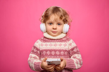 A young child with curly blonde hair wears white headphones and holds a smartphone. Dressed in a cozy pink sweater, the child appears absorbed in the music