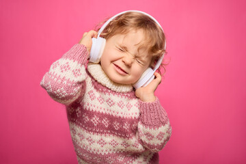 A young child with curly hair wears oversized headphones, smiling tightly with eyes shut in delight...