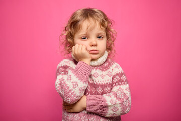 A toddler with curly hair stands with a bored expression. Wearing a cozy sweater, she rests her chin on her hand while gazing thoughtfully at the camera, set against a vibrant pink backdrop