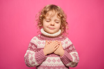 A young child with curly hair wears a cozy sweater and smiles softly while placing hands over their heart. The vibrant pink background enhances the warm atmosphere