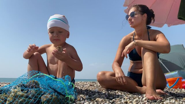 Beach day with mom and net full of seashells. Young boy sits on the beach wearing colorful striped hat and looking down at net full of seashells. Sky is clear and blue, water in background is light bl