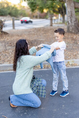 In a serene park setting filled with the beauty of nature, a caring mother lovingly assists her young son in putting on his jacket, capturing a precious moment of warmth and connection between them