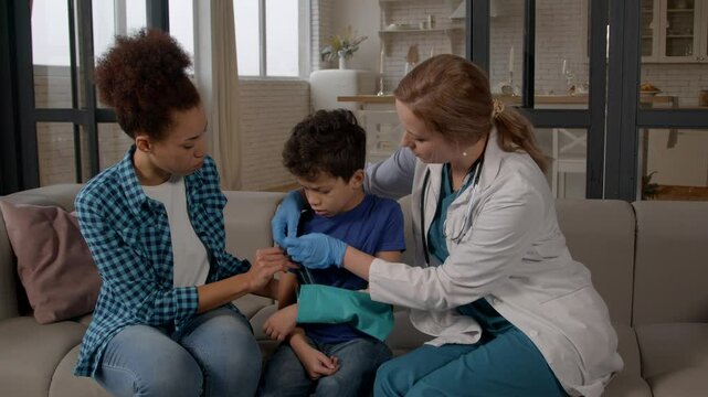 Caring woman doctor in uniform with worried African American mother providing medical first aid, applying arm sling bandage to fracture of sad teenage black boy in domestic room.