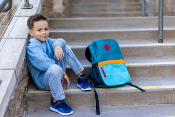 A cheerful young boy sits on steps next to a vibrant backpack, radiating joy and confidence. His...