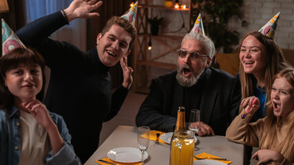 Family sit at table at home, grandchildren and parents celebrating birthday of grandfather, family members taking selfie on smartphone and smiling.