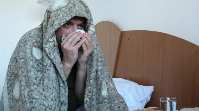 Unhappy sick mature man sitting on bed at home and treats flu and colds with medicinal drink. Middle aged shivering man wrapped in warm plaid drinking hot tea