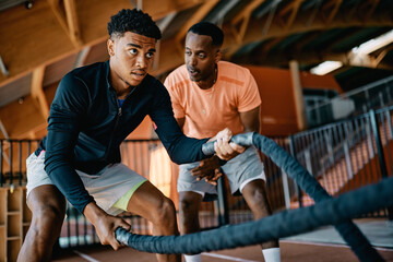 Instructor giving encouragement to a fit young man swinging battle ropes during a strength training session in a gym