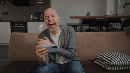 A middle-aged man at home on the sofa, laughing out loud while watching very funny videos on his smartphone. He s enjoying entertainment content from social media, immersed in the fun of the internet.