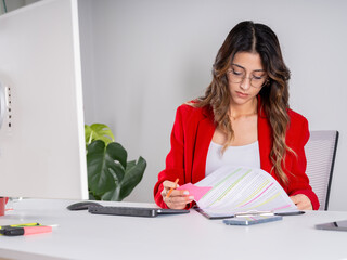 Working woman, front view young caucasian brunette attractive working woman sit desk office wear red suit formal jacket reading contract papers getting ready for meeting with client.