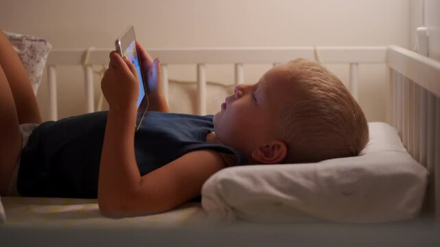 Toddler using tablet in bed late night tech addiction. Young boy lying in bed facing a tablet device. Child eyes are looking intently at screen of tablet. Little boy face is lit up by the screen.
