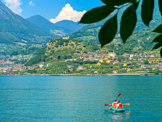 This beautiful image shows people kayaking on a peaceful lake Iseo, surrounded by breathtaking mountains and lush greenery. The calm waters reflect the vibrant colors of nature. Summer in Italy.
