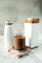A glass of cocoa protein milkshake surrounded by a milk carton and a plastic bottle.
