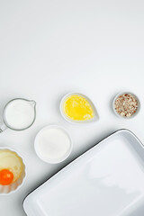 Flat lay composition for cooking preparation. Empty white baking dish with ingredients for baking, including egg, milk, butter, sugar, and nuts