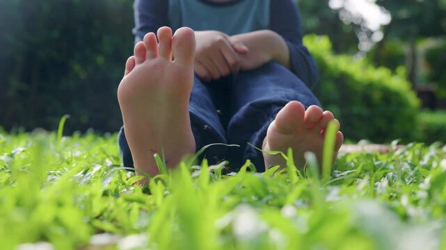 A young boy sitting on the grass wiggles his toes to shoo away mosquitoes that have landed on them