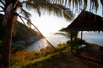 A serene coastal scene at sunset with a palm tree and a rustic gazebo overlooking the ocean.