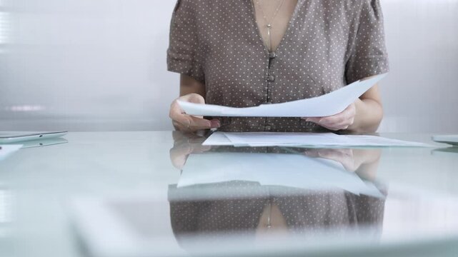 Businesswoman or accountant in brawn dotted blouse is going through paperwork while holding documents at a glass desk in a well lit, contemporary office setting. Business people concept