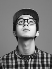 Fototapeta premium Studio portrait of young Asian male construction worker in black and white, wearing eyeglasses and cap, gazing upwards against gray backdrop, ample copyspace.