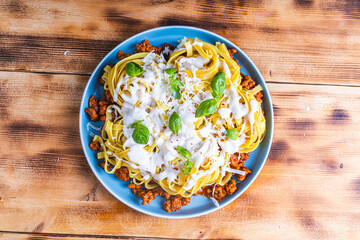 Spaghetti Bolognese pasta nests with Bechamel sauce and minced beef fried with tomatoes, onions and carrots on a blue plate on a wooden table. Top view. Copyspace or space for text.
